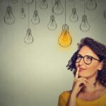 Thinking woman in glasses looking up with light idea bulb above head isolated on gray wall background